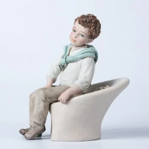 Figura in porcellana Alan, ragazzo seduto su poltrona bianca con camicia bianca e maglioncino azzurro sulle spalle.