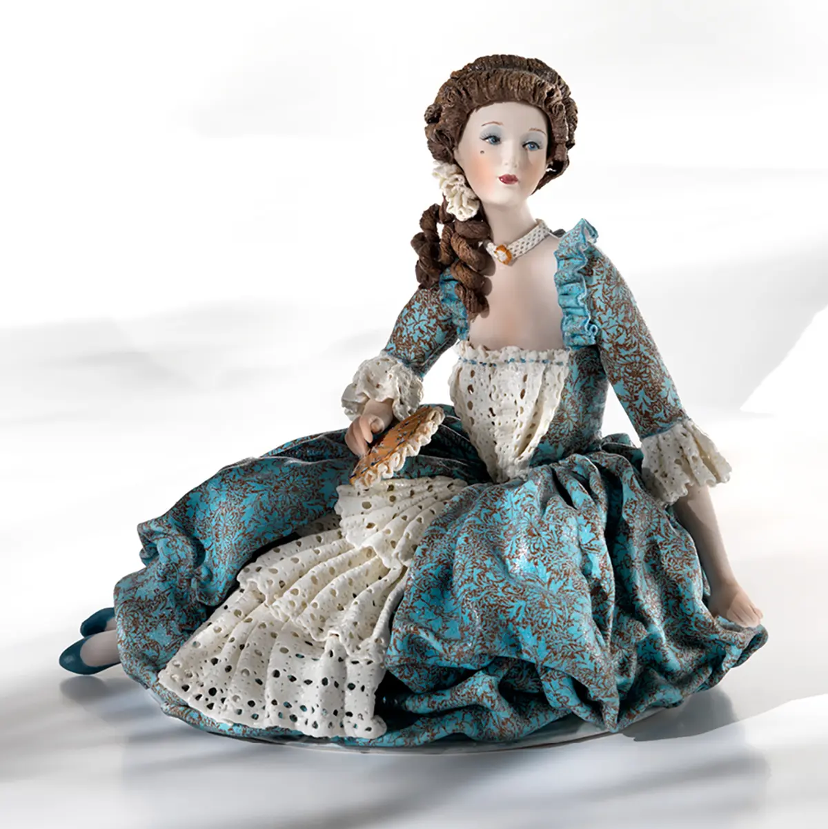 Statua in porcellana di dama veneziana del '700 seduta a terra, in abito azzurro con dettagli in pizzo e ventaglio ocra.