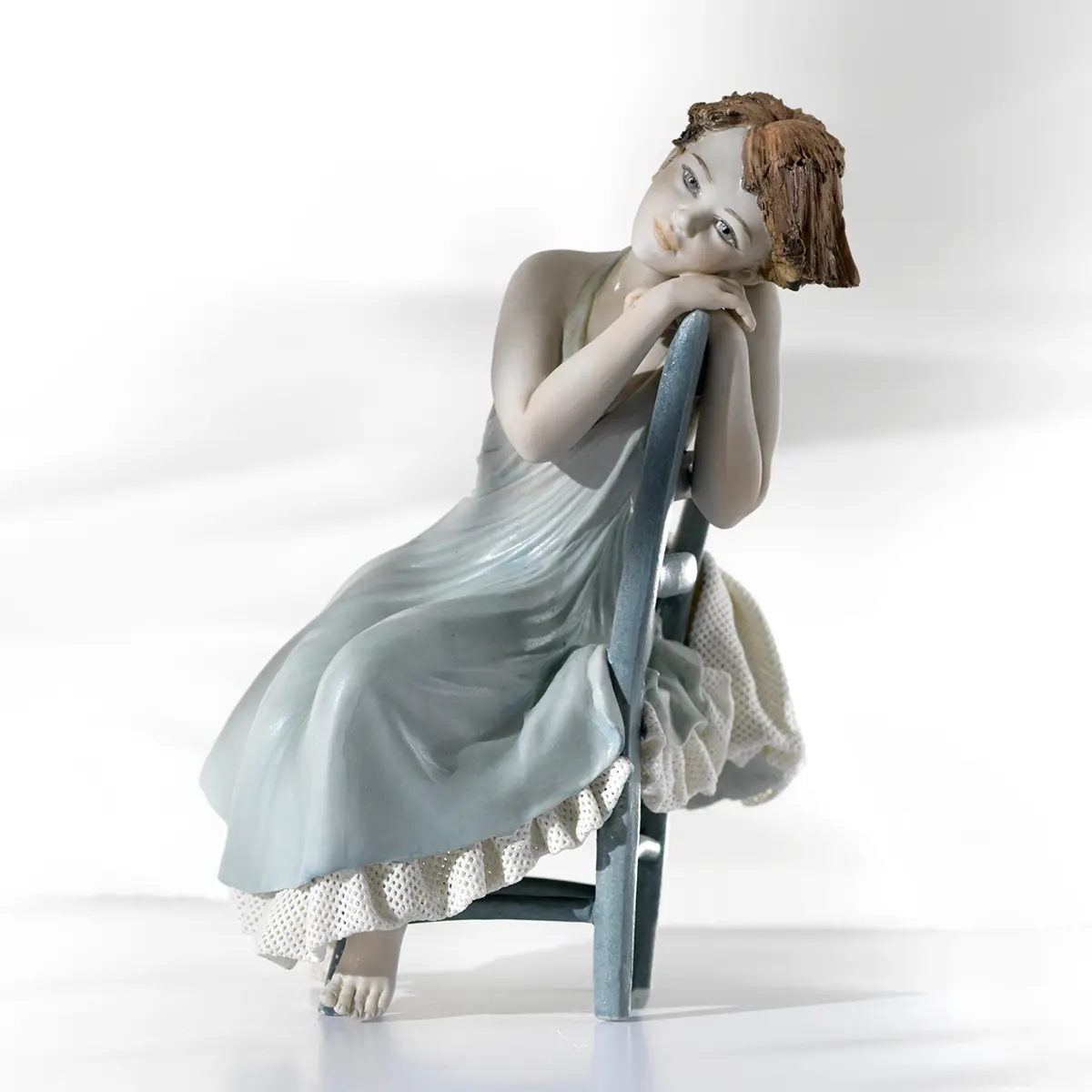 Statua in porcellana di giovane donna in abito celeste seduta su sedia turchese appoggiata allo schienale.
