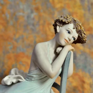 Statua in porcellana di giovane donna in abito azzurro seduta su una sedia turchese, appoggiata allo schienale.