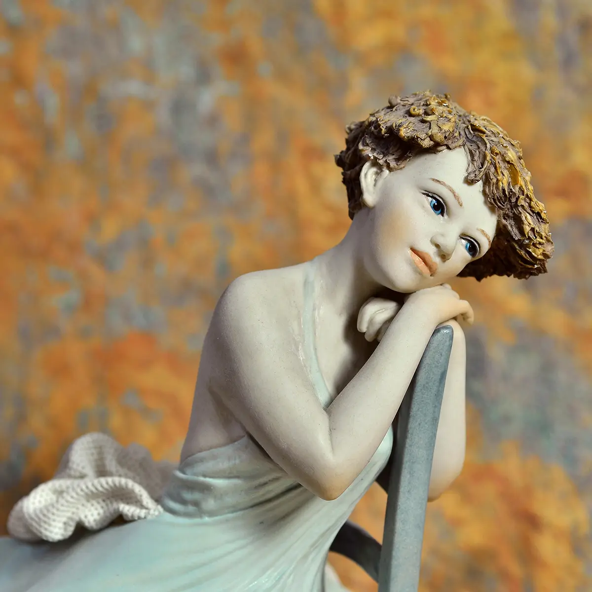 Statua in porcellana di giovane donna in abito azzurro seduta su una sedia turchese, appoggiata allo schienale.