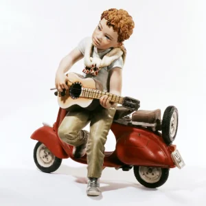 Statua in porcellana di ragazzo con chitarra appoggiato a una Vespa rossa, con t-shirt azzurra e pantaloni color senape.