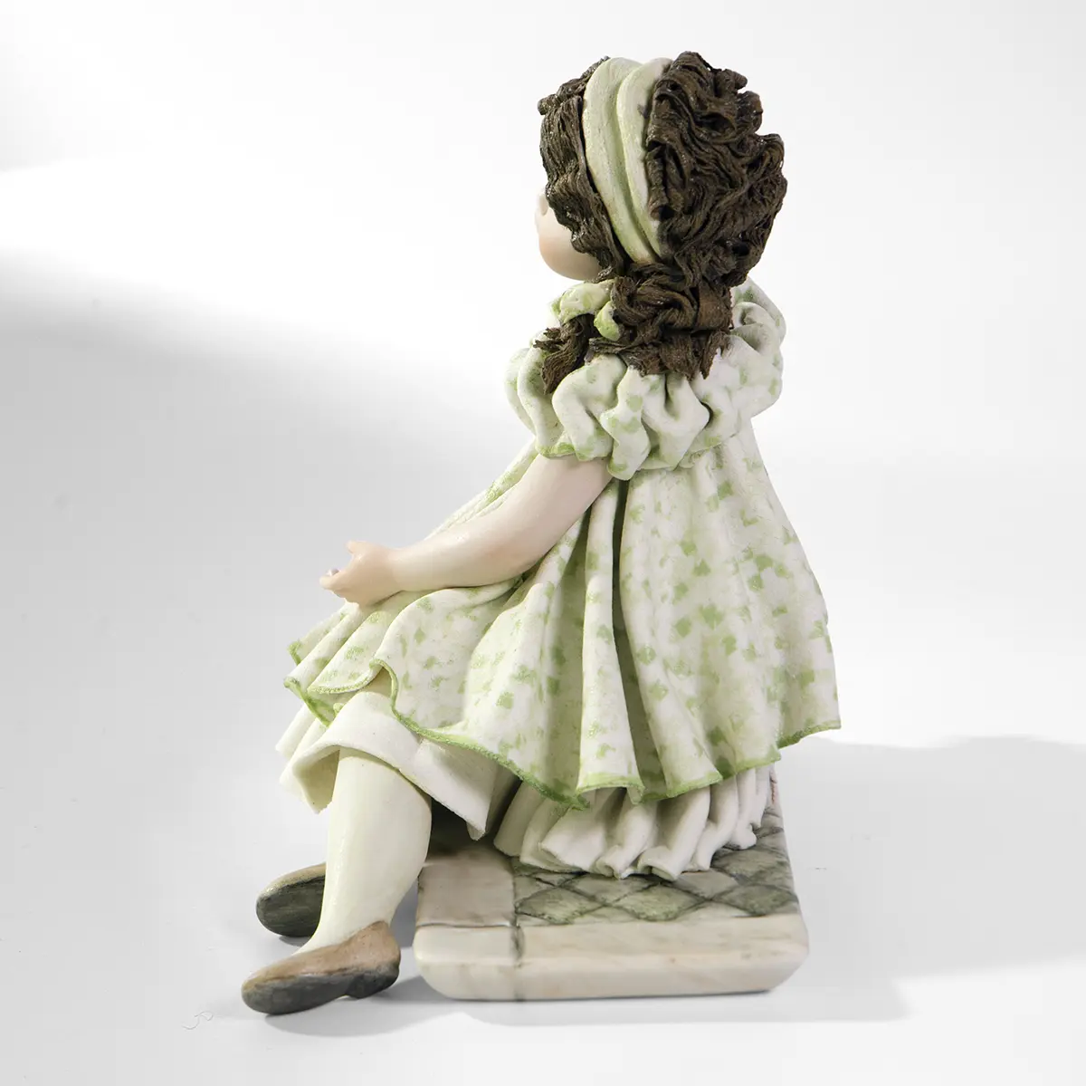 Statua in porcellana di ragazza con abito verde pastello e fascia bianca nei capelli, seduta su uno scalino marmorizzato.