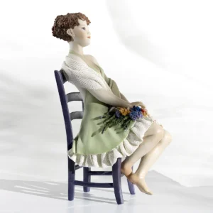 Statua in porcellana raffigurante donna in abito verde pastello seduta su sedia glicine con un mazzo di fiori colorati.