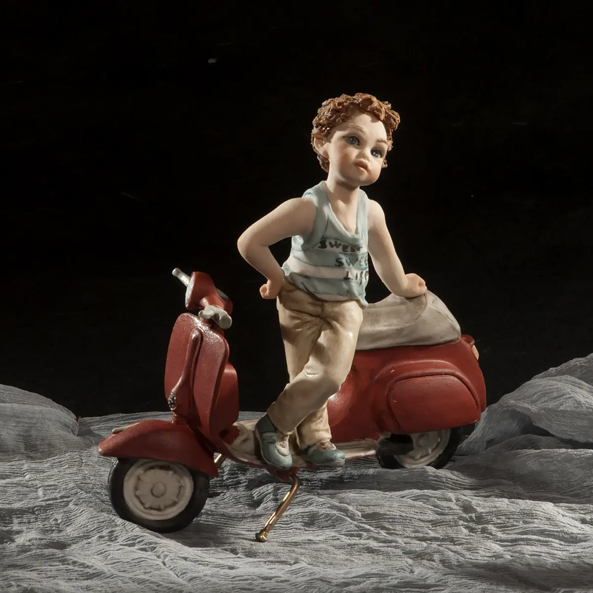 Statua in porcellana di ragazzino in piedi su Vespa rossa, con canottiera a righe bianche e azzurre e pantaloni beige