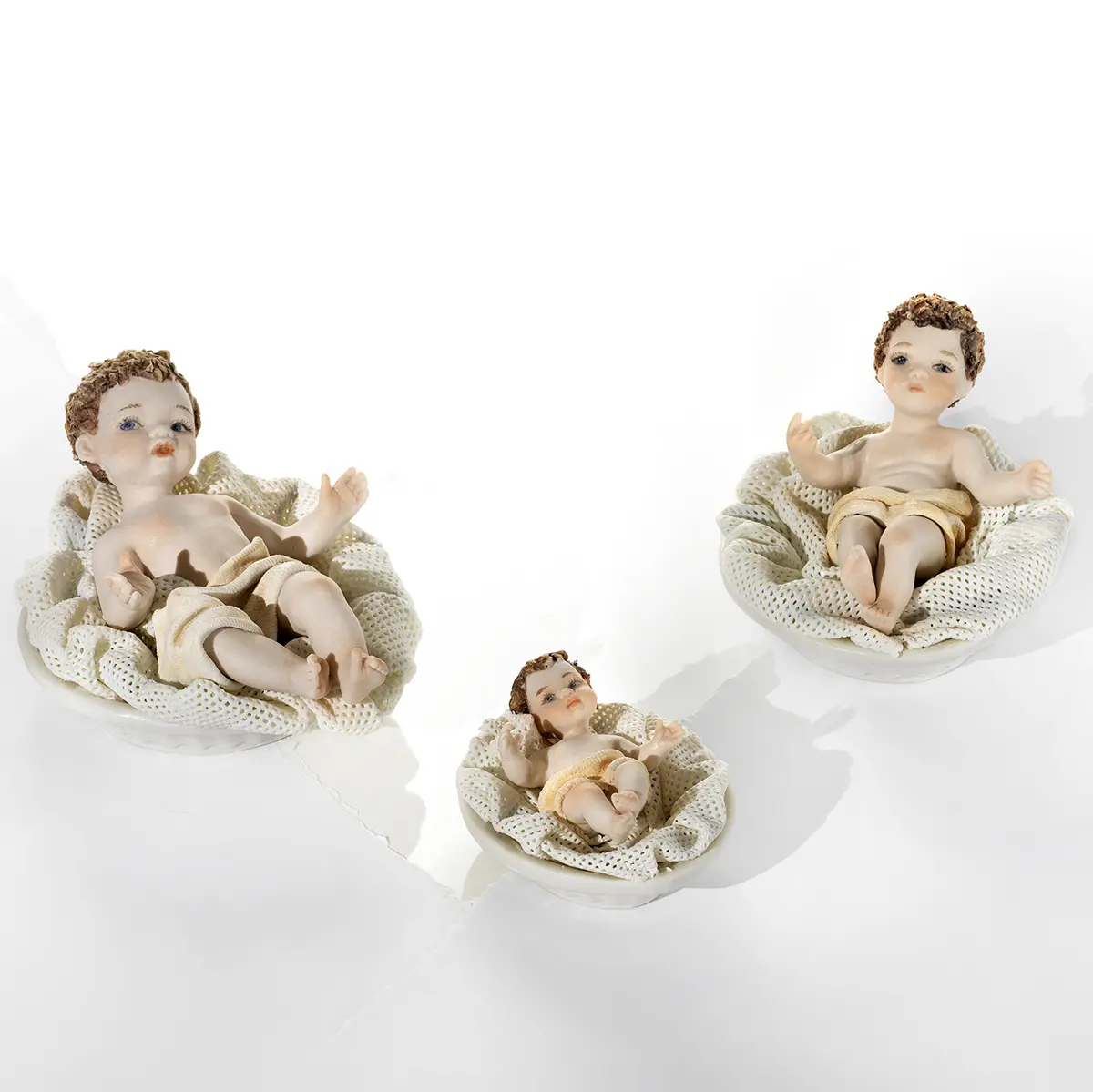 Tre statue in porcellana di Gesù Bambino in culla di diverse dimensioni, con panno color crema e cuscino in tulle.