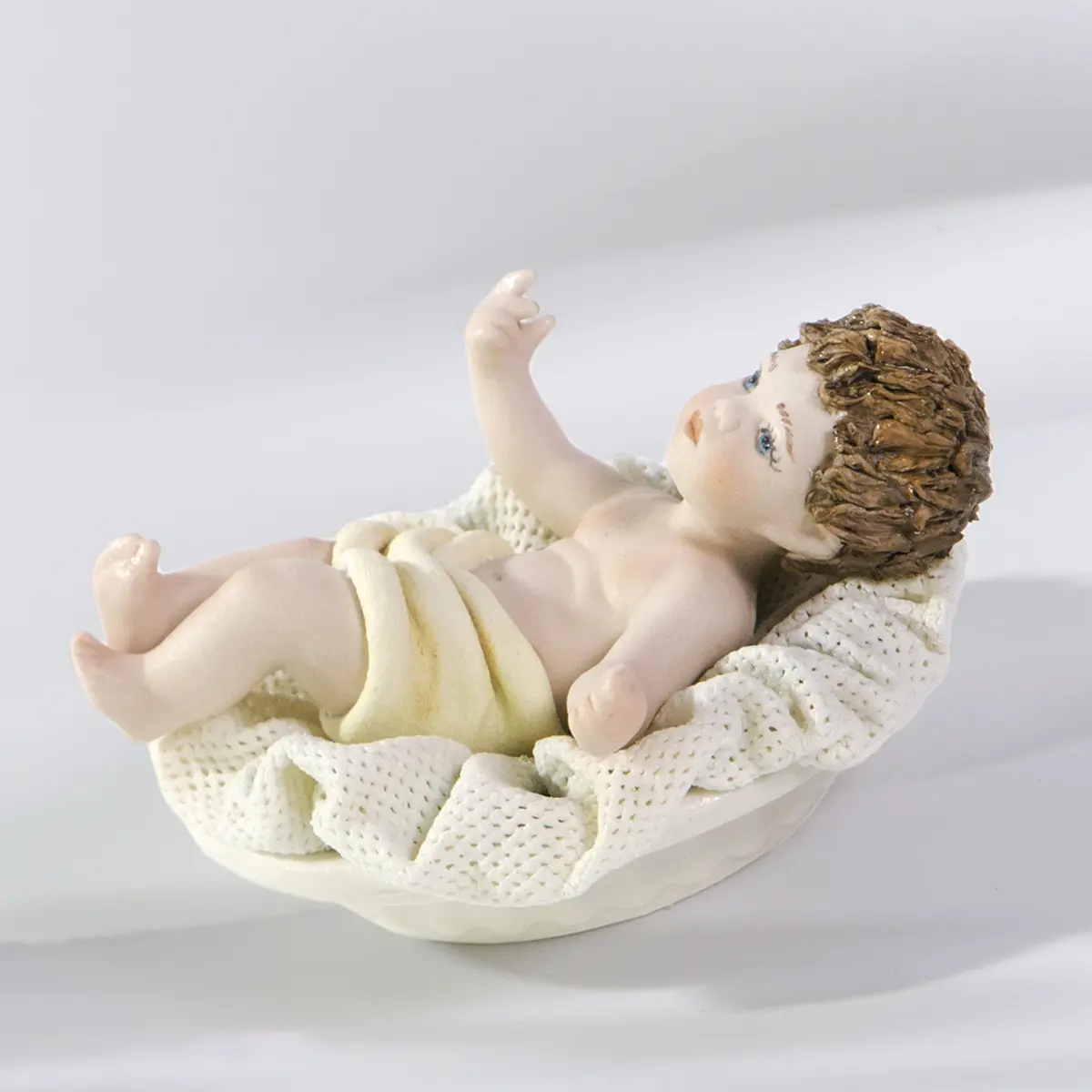 Figura in porcellana di Gesù Bambino con panno crema, steso su una culla con cuscino in tulle.