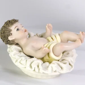 Statua in porcellana di Gesù Bambino steso su una culla con cuscino in tulle e panno crema.