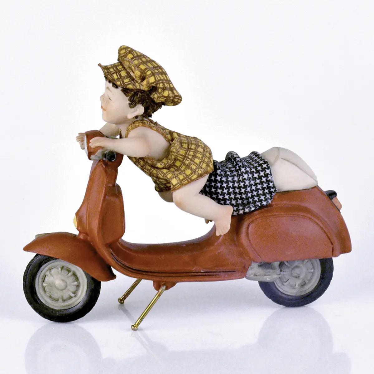 Statua in porcellana con bimbo sulla Vespa rossa, in salopette e berretto pied de poule giallo e marrone.