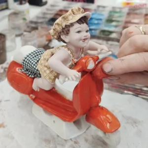 Statua in porcellana con bimbo su Vespa rossa, salopette e berretto pied de poule giallo e marrone.