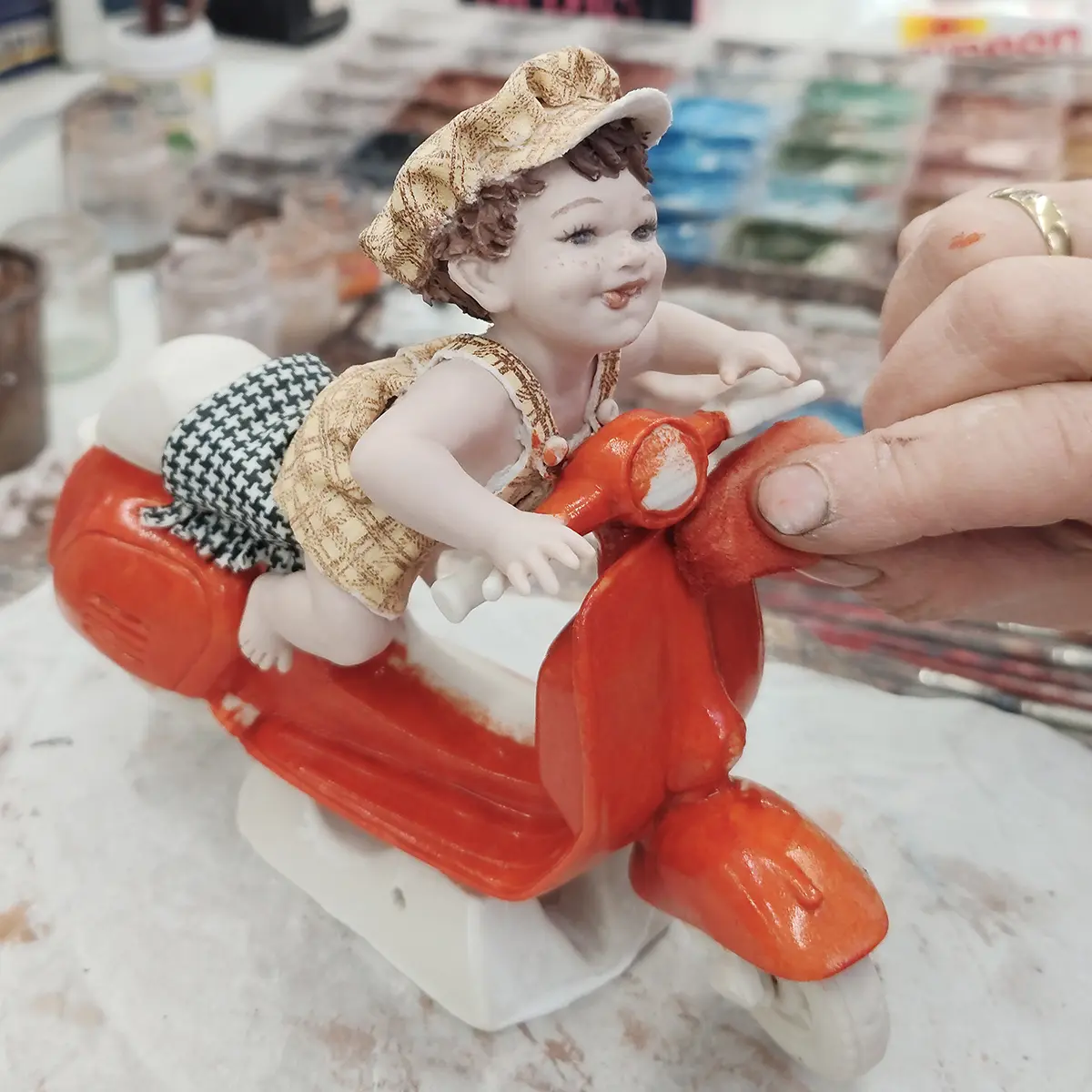 Statua in porcellana con bimbo su Vespa rossa, salopette e berretto pied de poule giallo e marrone.