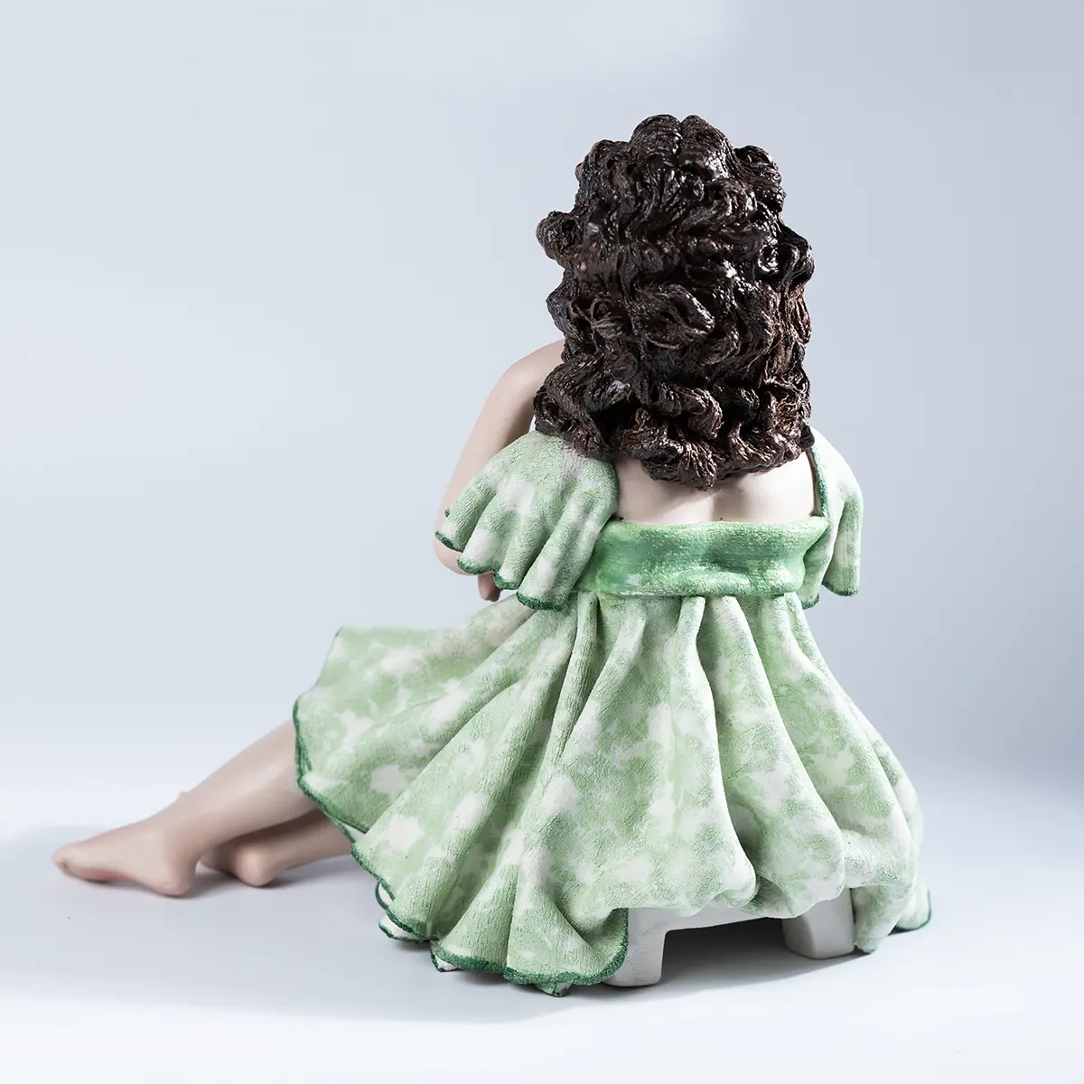 Statua in porcellana di ragazza seduta su sgabello, con abito verde tè, bordi verde smeraldo e capelli sciolti.