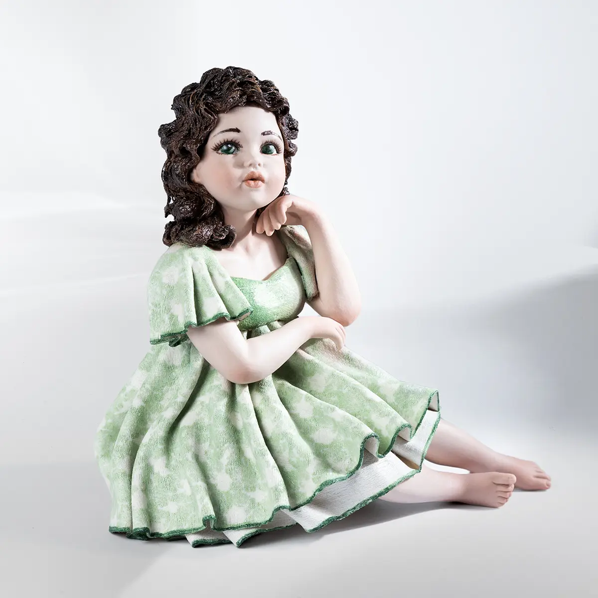 Statua in porcellana di ragazza seduta su sgabello, con abito verde tè, bordi smeraldo e capelli castani sciolti.