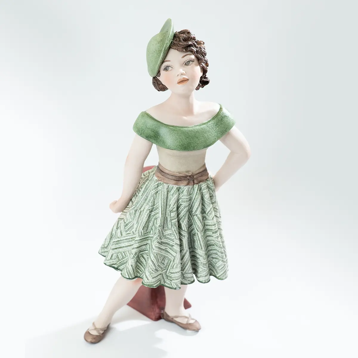 Statua in porcellana di Maggie con gonna verde e bianca, bustino beige e cappellino fascinator appoggiata a un tavolino.