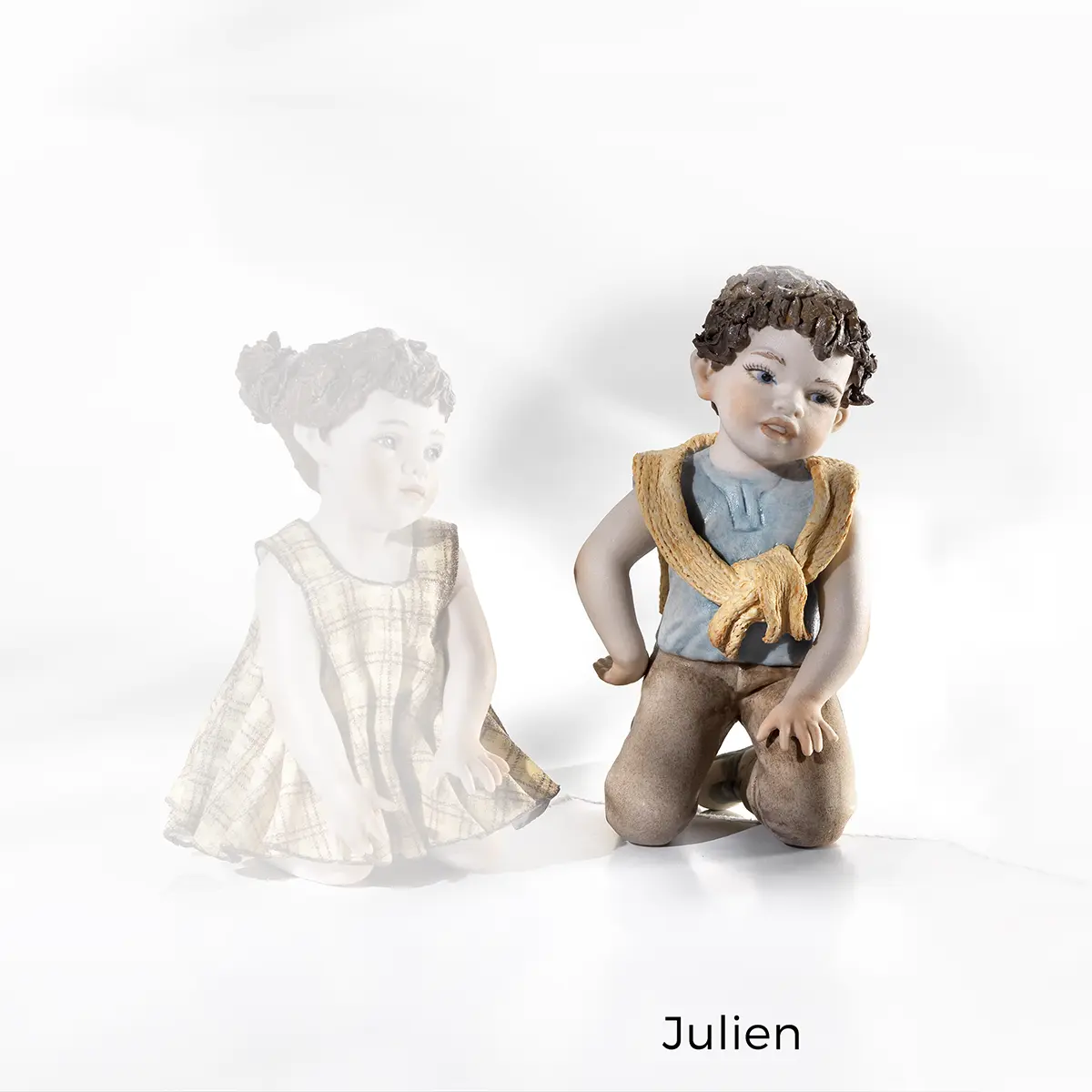 Statua in porcellana Julien con bimbo in ginocchio, t-shirt azzurra, maglioncino giallo sulle spalle e pantaloni marroni.