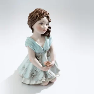 Statua in porcellana di mamma in ginocchio con abito celeste e fiore in mano, capelli castani sciolti su una spalla.