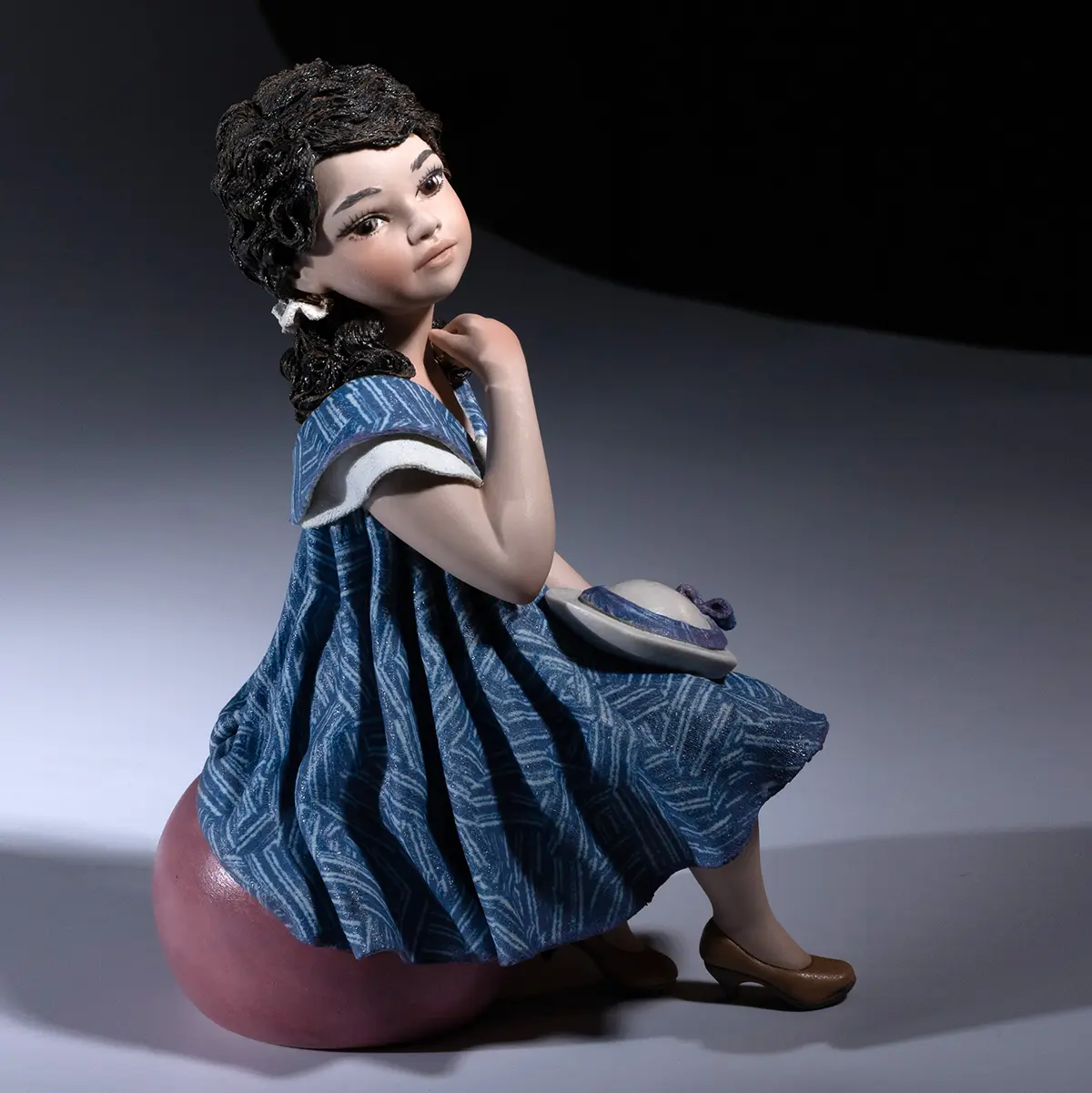 Statua in porcellana Nadia 2025 con abito indaco, seduta su pouf malva e cappellino bianco in grembo.