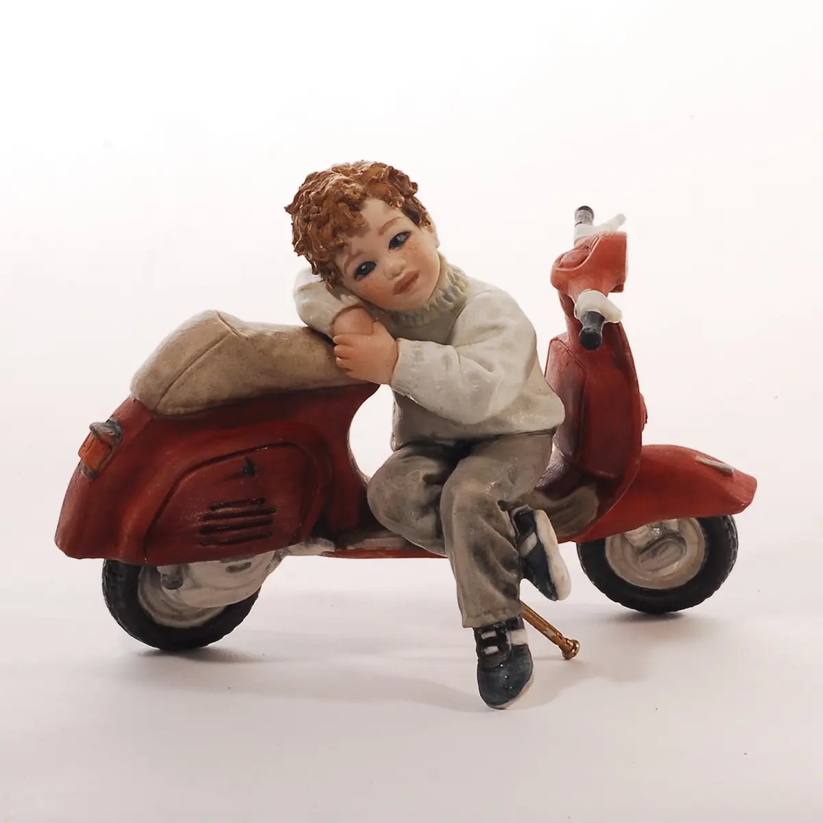 Statua in porcellana di un ragazzino seduto su una Vespa rossa con maglioncino azzurro e pantaloni grigi.
