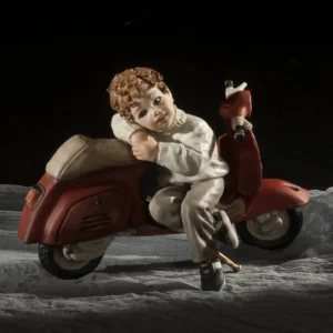 Statua in porcellana di ragazzino appoggiato a una Vespa rossa, con maglioncino azzurro chiaro e pantaloni grigi.