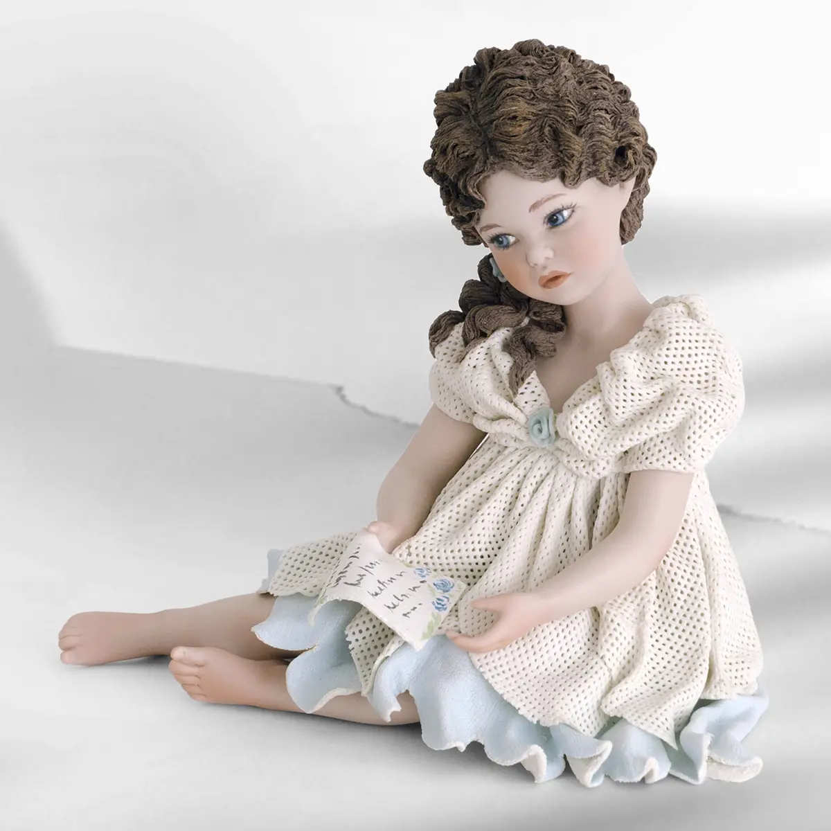 Statua in porcellana di ragazza seduta con abito bianco e azzurro, riccioli sulla spalla e una lettera in mano.