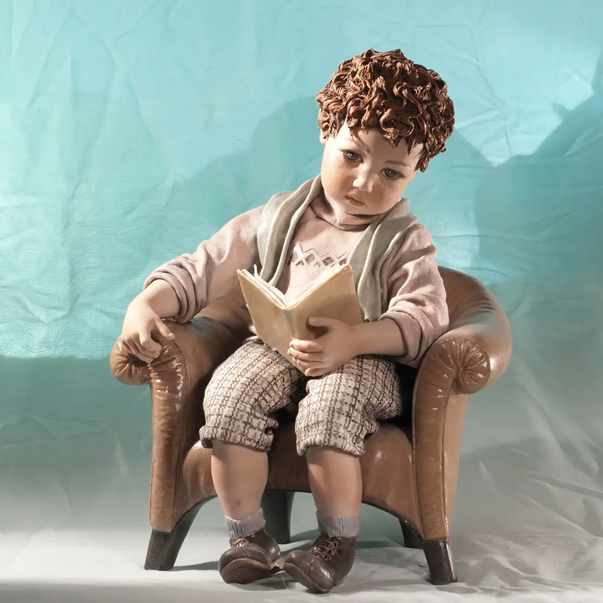 Statua in porcellana con bimbo seduto su poltrona, maglioncino salmone a rombi e libro di poesie aperto sulle gambe.